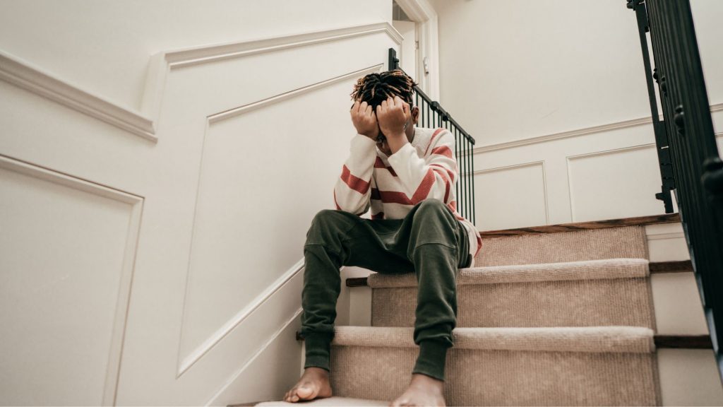 Contextual safeguarding, image of upset young male sat on the stairs, hands covering his face.