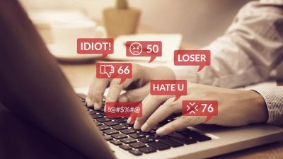 An image representing cyberbullying with a person on a laptop but with lots of message popping up saying horrible things like Loser, Idiot etc.