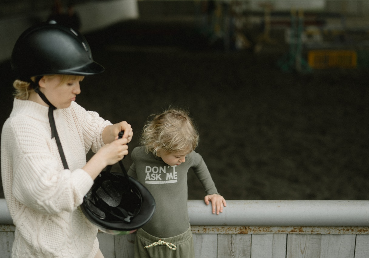 Woman fastening a riding helmet beside a young child at an indoor riding arena